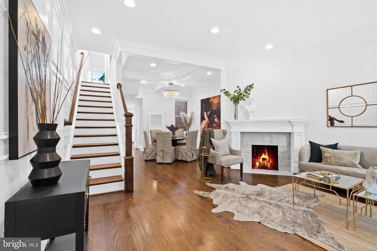The interior of a townhouse with white walls, wood floor, stairs on the left, a fireplace on the right, upscale furniture